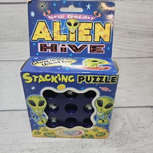 May include: A blue cardboard box for the New Galaxy Alien Hive stacking puzzle. The packaging features yellow alien graphics, the text "Alien Hive" and "Stacking Puzzle," and a cutout window revealing small, round, white and black alien game pieces.