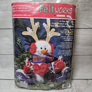 May include: A Feltworks craft kit for a snowman, featuring a snowman with antlers, earmuffs, and a scarf. The kit includes materials for stitching and glue assembly. The package is approximately 13 inches high.