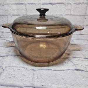 May include: A transparent amber-colored glass casserole dish with a matching lid. The dish has two side handles and a dark brown knob on the lid. The dish is designed for cooking and serving food.