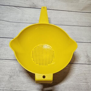 May include: A bright yellow plastic colander with a handle and a perforated base. The colander has a rounded shape with a handle extending from the top and a rectangular base. The colander is designed for draining liquids from food.