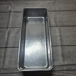May include: A rectangular silver-colored metal loaf pan with a smooth interior surface and slightly rounded corners. The pan features a metallic finish with visible light surface scratches and a flat bottom, designed for baking bread or cakes.