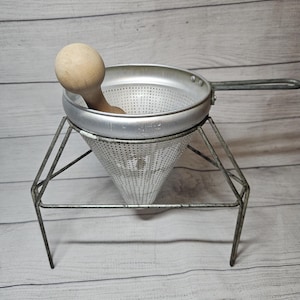 May include: A vintage metal food mill with a wooden pestle. The mill has a perforated bowl and a conical strainer, supported by a wireframe stand. The handle is attached to the side of the bowl. The wooden pestle rests inside the bowl.