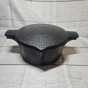 May include: A black kitchen pot with a matching lid featuring an integrated strainer section. The pot has two side handles and a pouring spout. The lid is circular with a textured surface and a series of small drainage holes for straining liquids.
