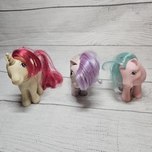 May include: Three small plastic toy ponies standing in a row. The first is a cream-colored unicorn with a bright pink mane and tail. The second is a light purple pony with a lavender mane. The third is a light pink pony with a mint green mane.