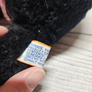 May include: A black plush toy with a pink and white patterned bow. A white tag with blue text reads "MINNIE MOUSE PRODUCT OF CONTENTS: SHREDDED CELL GROUND NUTS &copy; WALT DISN".