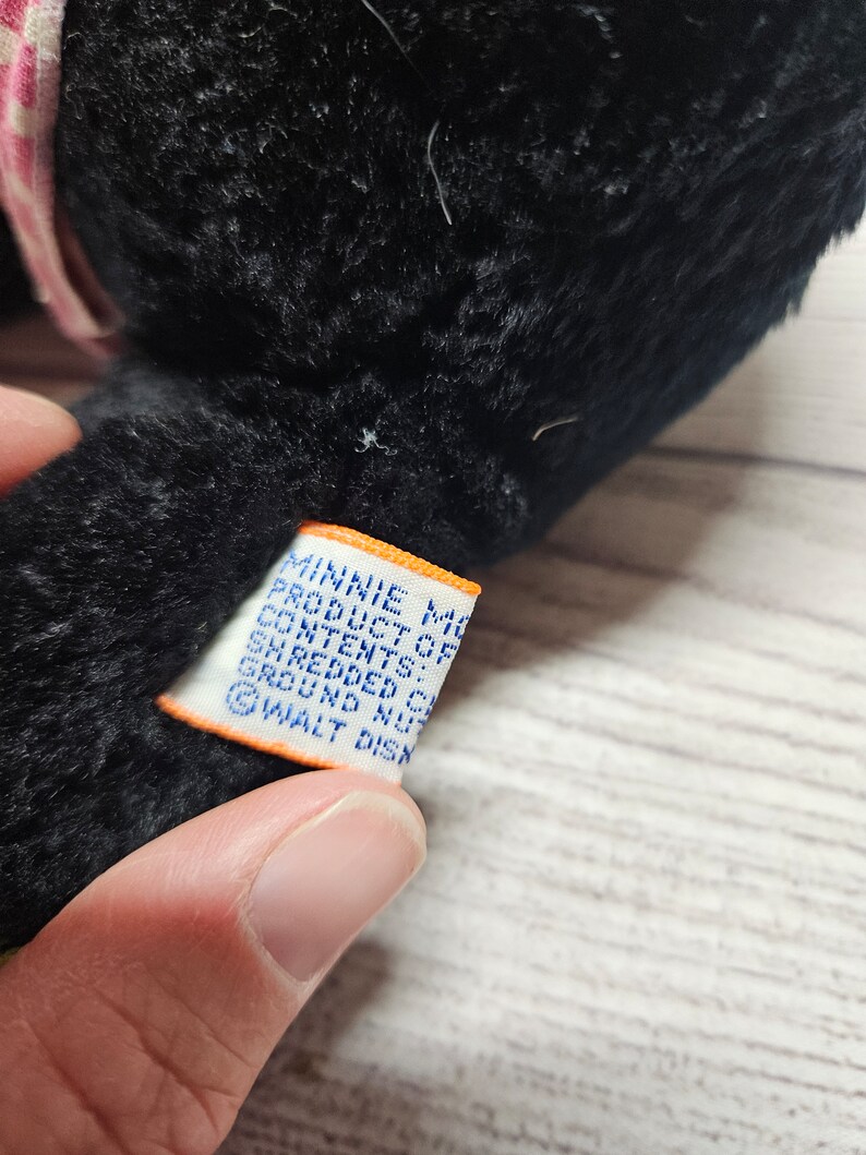 May include: A black plush toy with a pink and white patterned bow. A white tag with blue text reads "MINNIE MOUSE PRODUCT OF CONTENTS: SHREDDED CELL GROUND NUTS &copy; WALT DISN".
