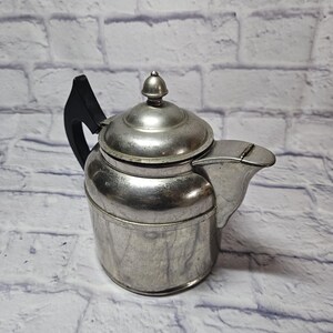 May include: A vintage silver-toned teapot with a black handle and a small, rounded lid. The teapot has a spout and a circular base. The background is a white brick wall.