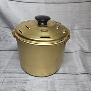 May include: A gold-colored metal cooking pot with a matching lid featuring a black knob handle. The lid has several small rectangular ventilation slots arranged in a circular pattern and a small arrow indicator pointing toward the edge.