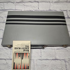May include: A gray backgammon set with three black stripes and a book titled "The Backgammon Book" by Oswald Jacoby and John R. Crawford. The book shows a backgammon board with red, black, and white pieces. The set is on a white brick background.