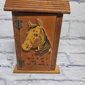 May include: A small, wooden cabinet with a hinged door and a sloped roof. The cabinet features a horse head graphic and text. The cabinet is brown with metal hinges and a small knob. The cabinet sits on a rectangular base.