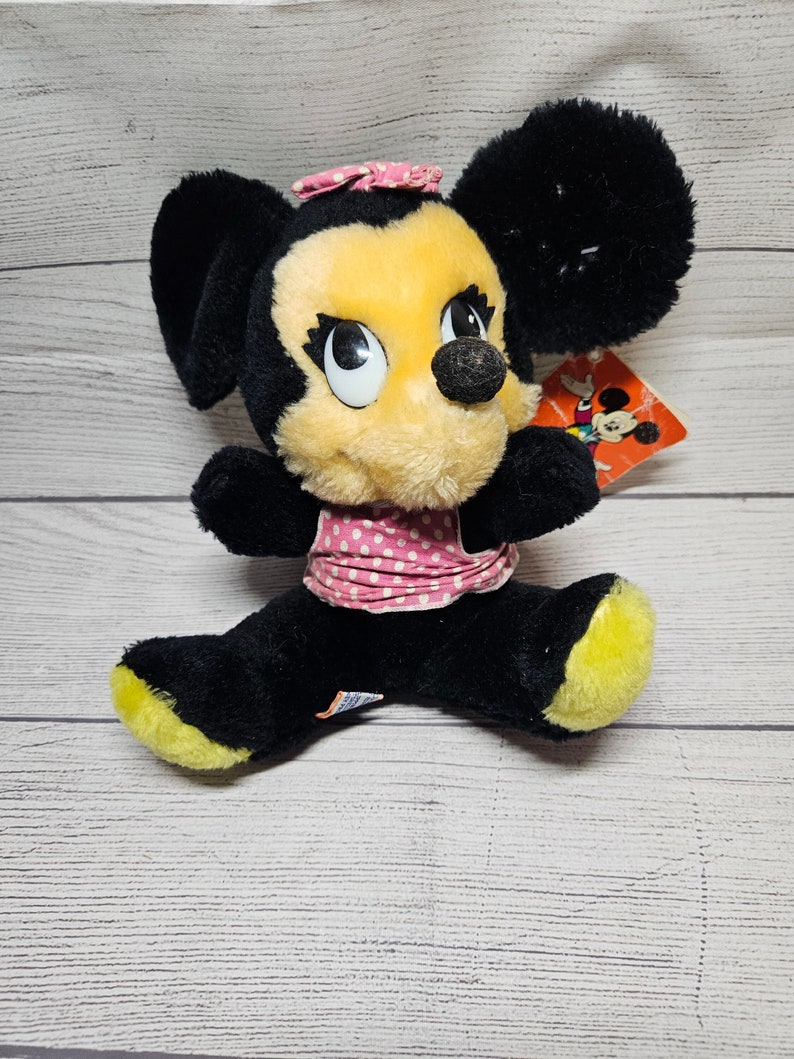 May include: Vintage Minnie Mouse plush toy. The plush features black fur, a yellow face, and a pink polka dot bow and dress. The toy has a tag attached. The plush is sitting on a white wooden surface.