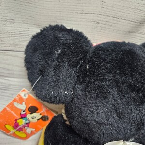 May include: A black plush Mickey Mouse toy with a white ribbon and a colorful tag featuring Mickey Mouse. The toy has large, round ears and a soft, textured appearance. The tag is orange with a cartoon image.