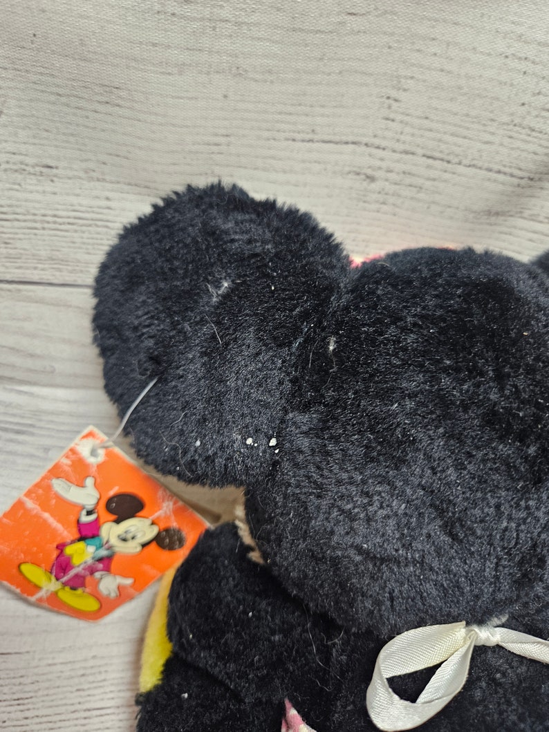 May include: A black plush Mickey Mouse toy with a white ribbon and a colorful tag featuring Mickey Mouse. The toy has large, round ears and a soft, textured appearance. The tag is orange with a cartoon image.