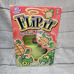 May include: A rectangular game box for Flip It The Topsy-Turvy Game featuring a red background with cartoon green turtles. The box displays the text 40 Challenge Cards and Ages 8 & Up. A cutout window reveals a light green plastic game component.