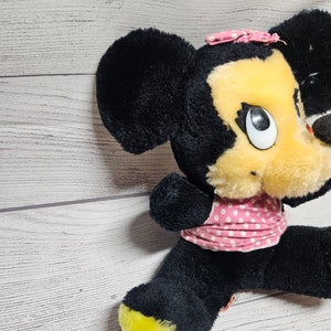 May include: A vintage plush toy of a cartoon mouse with black fur, a yellow face, and a pink polka-dot bow and dress. The mouse has large white eyes and a black nose. The feet are yellow.
