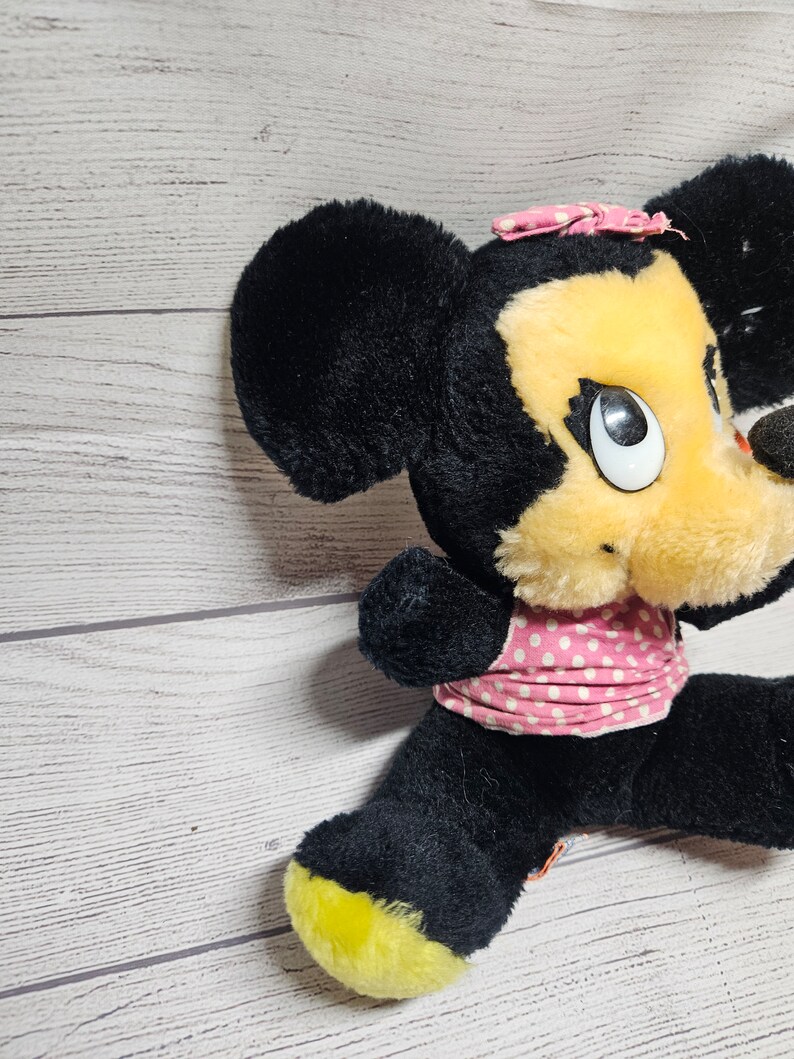 May include: A vintage plush toy of a cartoon mouse with black fur, a yellow face, and a pink polka-dot bow and dress. The mouse has large white eyes and a black nose. The feet are yellow.