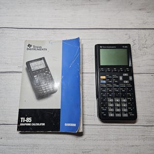 May include: A black TI-85 graphing calculator with a black and white guidebook. The calculator has a screen and numerous buttons. The guidebook is titled "TI-85 Graphing Calculator Guidebook" and features the Texas Instruments logo.