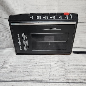May include: A black General Electric mini cassette recorder and player featuring a rectangular design with a clear window, a silver trim accent, and top-mounted control buttons labeled REC/BATT, RECORD, PLAY, REW/REV, F.FWD/PREV, STOP/EJECT, and PAUSE.
