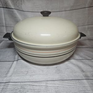 May include: A cream-colored round Dutch oven with a matching lid and black side handles. The pot features a black knob on the lid and horizontal decorative stripes in orange and dark brown around the exterior body, showcasing a classic kitchen design.