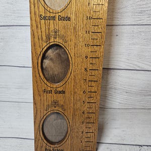 May include: Wooden growth chart with three oval photo frames labeled "Pre-School", "First Grade", and "Second Grade". A height scale in feet and inches is on the right side. The wood has a natural grain and a warm, brown tone.