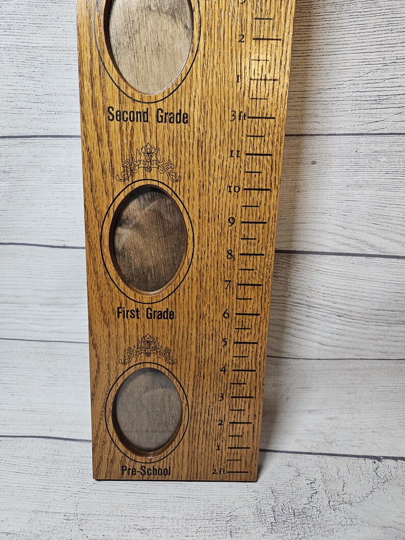 May include: Wooden growth chart with three oval photo frames labeled "Pre-School", "First Grade", and "Second Grade". A height scale in feet and inches is on the right side. The wood has a natural grain and a warm, brown tone.