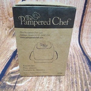May include: A cardboard box for a Pampered Chef food holder. The box is tan with black text and a logo. The text includes the company name, address, and product information. The box also has a line drawing of the food holder.