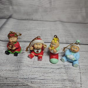 May include: Four small Cabbage Patch Kids ornaments with gold hanging loops. One child holds a brown teddy bear, one wears a Santa hat and red outfit, one sits in a red stocking, and one wears a blue outfit while holding a white snowball.