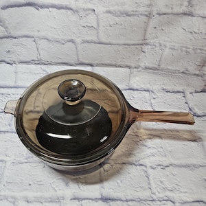 May include: A clear glass saucepan with a matching lid and a long handle. The cookware is a light brown color and sits on a white brick background. The lid has a small knob for easy handling.