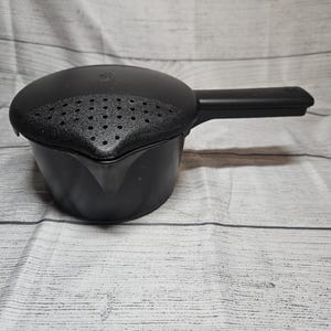 May include: A black kitchen saucepan with a matching lid featuring an integrated strainer. The lid has a textured surface with a series of small drainage holes near the spout area. The handle extends horizontally from the side of the round pot.