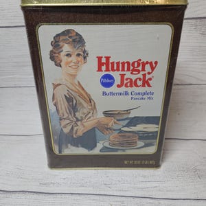 May include: A vintage Hungry Jack Buttermilk Complete Pancake Mix tin. The brown tin features a woman in a vintage dress and the Hungry Jack logo in red. The net weight is 32 oz (2 lb) 907g.