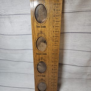 May include: Wooden growth chart with four oval photo frames for preschool, first, second, and third grade. The chart has a ruler on the side with measurements up to 11 inches. The wood has a natural grain and a warm, brown tone.