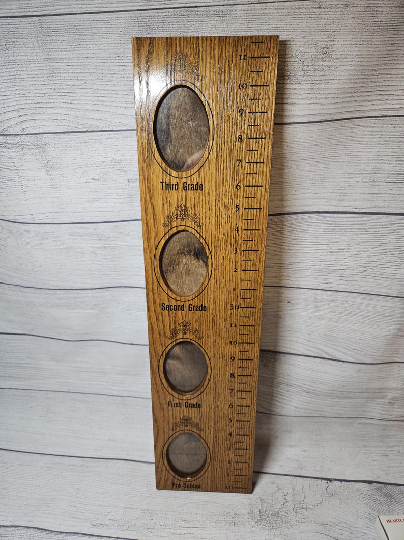 May include: Wooden growth chart with four oval photo frames for preschool, first, second, and third grade. The chart has a ruler on the side with measurements up to 11 inches. The wood has a natural grain and a warm, brown tone.