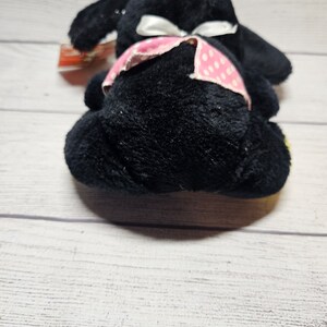 May include: A black plush toy bunny with floppy ears and a white bow. The bunny has a pink polka dot patterned band around its waist. The toy is lying on a light wood surface.