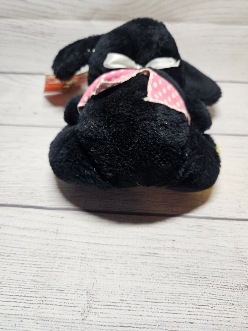 May include: A black plush toy bunny with floppy ears and a white bow. The bunny has a pink polka dot patterned band around its waist. The toy is lying on a light wood surface.
