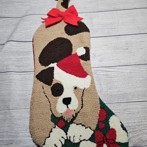 May include: A Christmas stocking featuring a dog design. The stocking has a tan background with brown spots, a red bow, and a green section with a red bow and white paws. The dog is wearing a Santa hat.