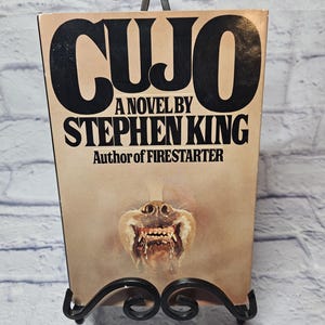May include: A vintage paperback book titled "CUJO" by Stephen King. The cover features the title in large black letters, the author's name, and a menacing illustration of a dog's snarling face. The book is displayed on a decorative black metal stand.