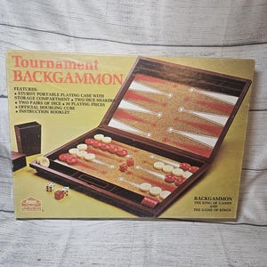 May include: A Tournament Backgammon board game box featuring a dark brown folding case with a cork-textured interior, red and white triangular points, thirty playing pieces, two dice shakers, and dice. The box includes text and game features.