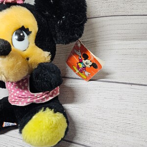 May include: A plush Minnie Mouse doll with black fur, a yellow face, and a pink polka-dot bow and scarf. The doll has a tag with a cartoon image of Mickey Mouse. The doll's feet are yellow.