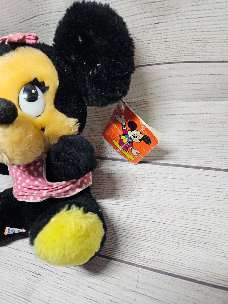 May include: A plush Minnie Mouse doll with black fur, a yellow face, and a pink polka-dot bow and scarf. The doll has a tag with a cartoon image of Mickey Mouse. The doll's feet are yellow.