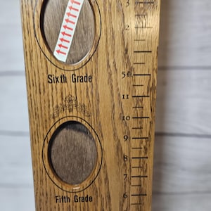 May include: Wooden growth chart with oval photo frames for Fifth and Sixth Grade. The chart has a ruler on the side with measurements in feet and inches. The chart is made of light brown wood.