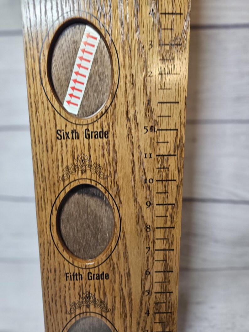 May include: Wooden growth chart with oval photo frames for Fifth and Sixth Grade. The chart has a ruler on the side with measurements in feet and inches. The chart is made of light brown wood.