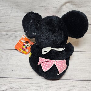 May include: A black plush toy mouse with large round ears and a pink polka-dot bow. The toy has a white bow at the neck and a colorful tag attached. The toy is sitting on a light-colored wooden surface.