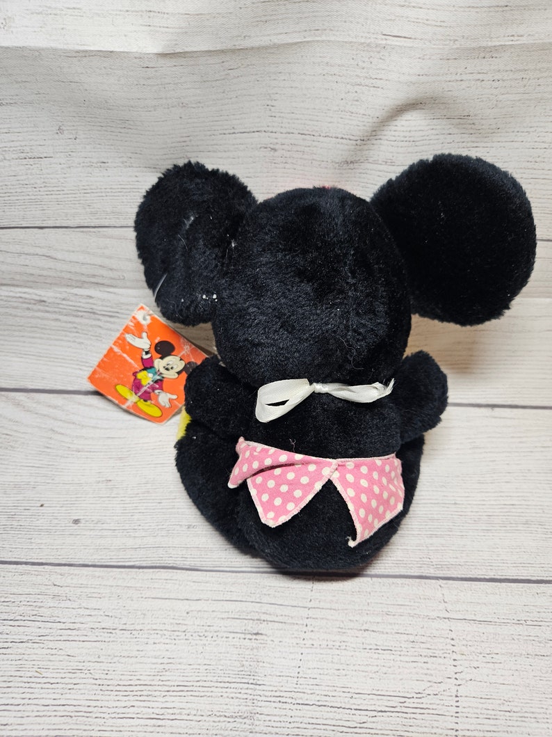 May include: A black plush toy mouse with large round ears and a pink polka-dot bow. The toy has a white bow at the neck and a colorful tag attached. The toy is sitting on a light-colored wooden surface.