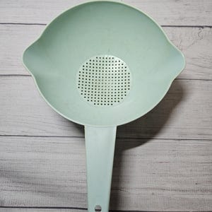 May include: A light blue plastic colander with a handle. The colander has a round bowl shape with a perforated bottom and two side handles. The handle has a hole at the end for hanging. The colander is designed for draining liquids from food.
