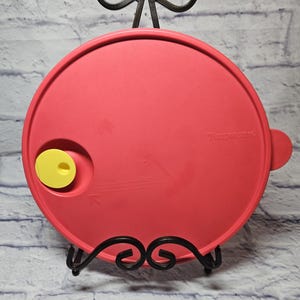 May include: A red Tupperware lid with a yellow valve and a handle. The lid is round and sits on a black metal display stand. The word "Tupperware" is printed on the lid.