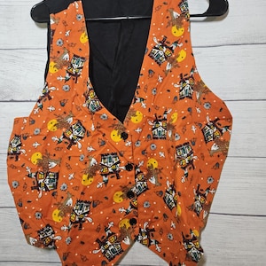 May include: A sleeveless vest featuring an orange fabric with a repeating Halloween pattern of haunted houses, yellow full moons, ghosts, and bare trees. The garment has a solid black back panel, a V-neckline, and four black buttons down the front.