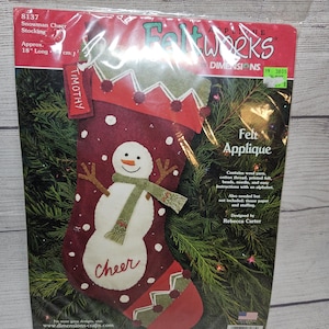 May include: A Christmas felt applique kit for a snowman stocking. The stocking is approximately 18 inches long and features a snowman with a green scarf and the word "Cheer". The kit includes wool yarn, cotton thread, and printed felt.
