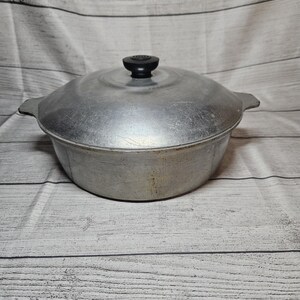 May include: A round, silver-colored aluminum pot with a matching lid and a small black knob handle on top. The pot features two integrated side handles and has a brushed metal finish with visible signs of wear and light surface scratches.
