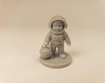 Puede incluir: Una pequeña figura gris monocromática de un niño vestido con un traje de astronauta detallado. La figura lleva un casco redondeado, un panel de control en el pecho y sostiene un pequeño cubo redondo mientras está de pie sobre una base circular.
