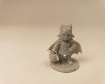 Puede incluir: Una pequeña figura de resina gris sin pintar de un personaje que lleva una máscara de murciélago, una capa y una túnica con un emblema de murciélago en el pecho. La figura se apoya sobre una base circular y sostiene un pequeño cubo redondo en una mano.